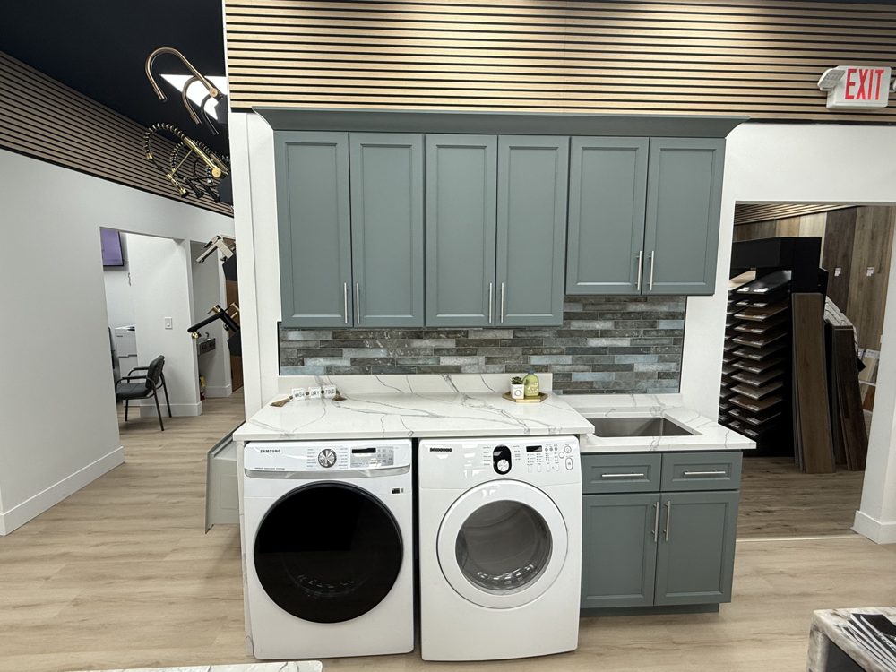Laundry room display with sage green shaker cabinets, a white veined countertop over the washer and dryer, a utility sink, and a grey brick-look backsplash
