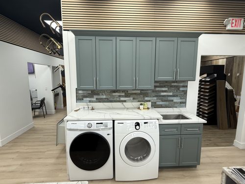 Laundry room display with sage green shaker cabinets, a white veined countertop over the washer and dryer, and a utility sink