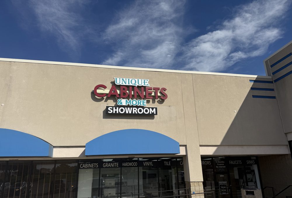 The Unique Cabinets & More showroom storefront on Haggerty Road, with its red and teal sign, blue awnings, and window lettering for cabinets, granite, flooring, faucets, and sinks